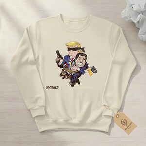 May include: Cream-colored sweatshirt featuring a cartoon illustration of two figures. One figure, with a blindfold and a gun, carries another figure tied up. The word "CAPTURED" is printed on the front.