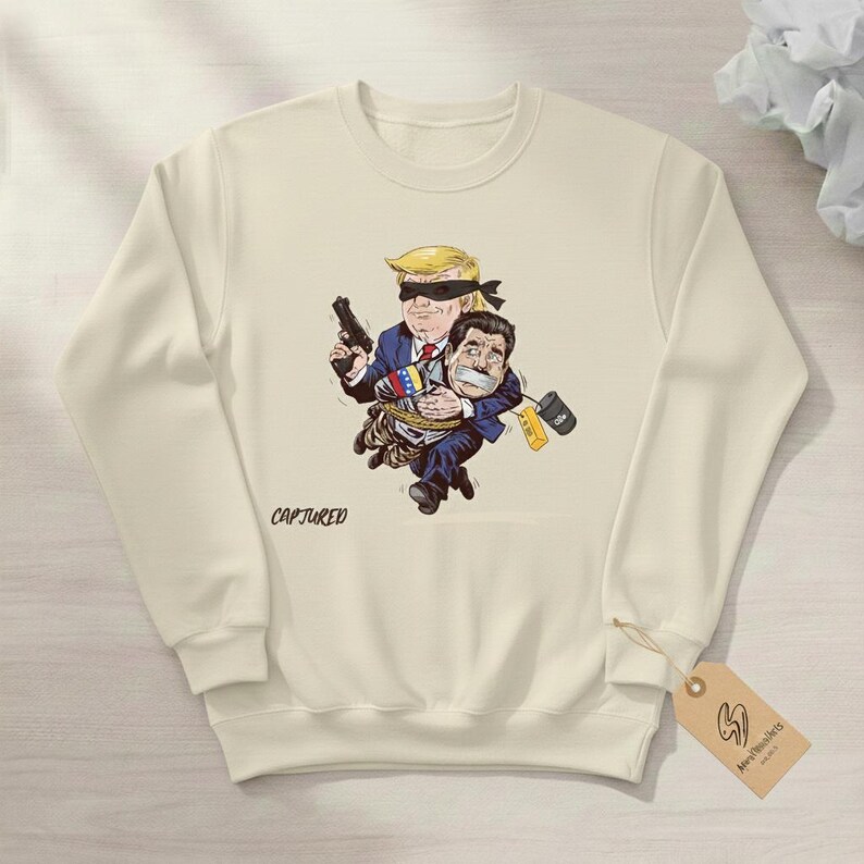 May include: Cream-colored sweatshirt featuring a cartoon illustration of two figures. One figure, with a blindfold and a gun, carries another figure tied up. The word "CAPTURED" is printed on the front.