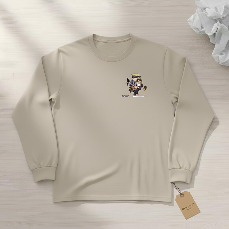 May include: A long-sleeved, beige shirt featuring a small cartoon graphic of two figures. One figure is holding a gun and the other is holding a small bag. A small tag is attached to the shirt.