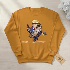 May include: Mustard yellow sweatshirt featuring a cartoon illustration of two figures. One figure, with a blindfold and a gun, is holding the other figure, who is gagged and bound. The word "CAPTURED" is printed on the front.