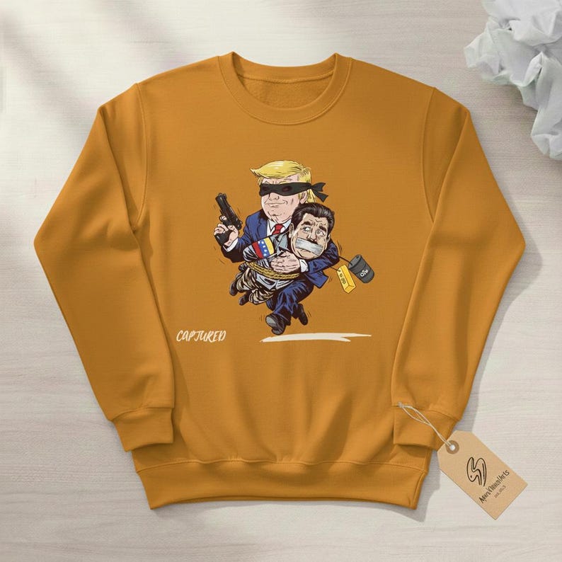 May include: Mustard yellow sweatshirt featuring a cartoon illustration of two figures. One figure, with a blindfold and a gun, is holding the other figure, who is gagged and bound. The word "CAPTURED" is printed on the front.