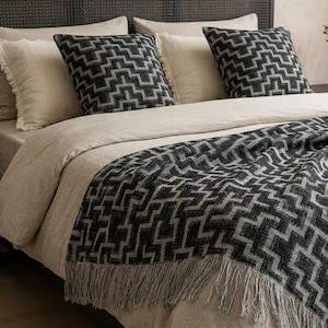 Può includere: Un letto con biancheria da letto beige, due cuscini color crema e due cuscini quadrati con un motivo geometrico nero e grigio. Una coperta abbinata con frange è drappeggiata ai piedi del letto.
