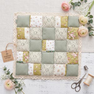 Peut inclure: Un quilt patchwork carré composé d'une grille de carrés en tissu alternant vichy vert, motifs floraux et dentelle crème. Le quilt est bordé d'une garniture en dentelle festonnée et comprend une étiquette brune marquée HANDMADE COTTAGE.