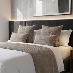 May include: A bed with white linens, a textured beige bed runner, and matching pillows. The headboard is dark gray, and a framed artwork hangs above the bed. A small wooden nightstand with a lamp is on the right.