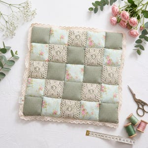 May include: A square patchwork quilt featuring a grid of alternating fabric squares in green gingham, light blue floral print, and cream lace. The quilt is edged with a cream lace trim and measures approximately 10 inches by 10 inches.