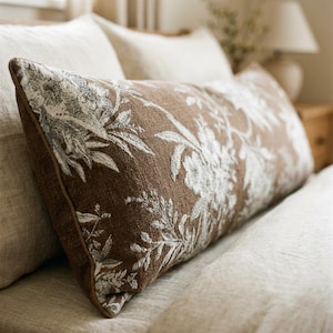 May include: A rectangular brown pillow with a white floral pattern. The pillow has a brown border and is placed on a bed with white and beige bedding. The pillow's design features detailed botanical illustrations.