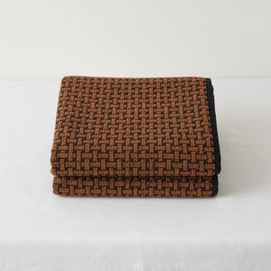 May include: Folded brown and black woven textured towels. The towels are stacked neatly, showcasing a detailed basketweave pattern. The edges are finished with a solid black border, creating a contrasting visual element. The towels are on a white surface.