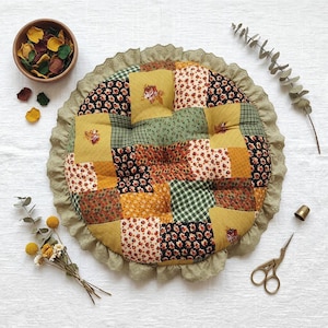 May include: A round patchwork cushion featuring a variety of fabric squares in mustard yellow, forest green, and black with floral and gingham patterns. The cushion is finished with a wide, scalloped sage green fabric ruffle border.