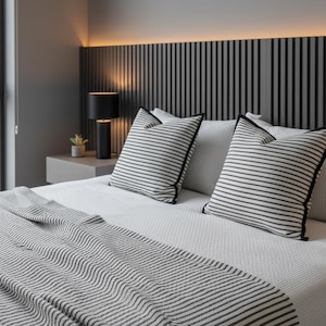 May include: A modern bedroom scene featuring a bed with white bedding, striped pillows with black trim, and a matching throw blanket. A black lamp sits on a floating nightstand, and a decorative headboard with vertical black and gray stripes completes the look.