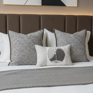 Könnte beinhalten: Ein Bett mit mehreren dekorativen Kissen und einem Bettläufer. Die Kissen sind grau mit braunen Rändern, und ein kleineres Kissen zeigt ein kreisförmiges Design. Der Bettläufer ist grau, und das Kopfteil ist braun.