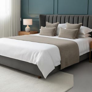 Könnte beinhalten: Ein modernes Schlafzimmer-Setup mit einem grau gepolsterten Bett und einem hohen Kopfteil. Das Bett ist mit weißer Bettwäsche bezogen, akzentuiert durch beige Kissen und einen passenden Bettläufer. Ein kleiner Nachttisch aus Holz mit einer Lampe steht neben dem Bett, und ein weißer Teppich liegt auf dem Boden.