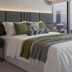May include: A contemporary bed featuring a grey upholstered headboard, white bedding, and decorative cushions in green and black and white patterns. A green throw and a grey fringed blanket add texture and colour.