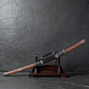May include: A Japanese katana sword with a wooden handle and scabbard, displayed on a wooden stand. The blade is partially visible, with a blue and black wrapped hilt. Silver-coloured metal accents adorn the handle and scabbard.