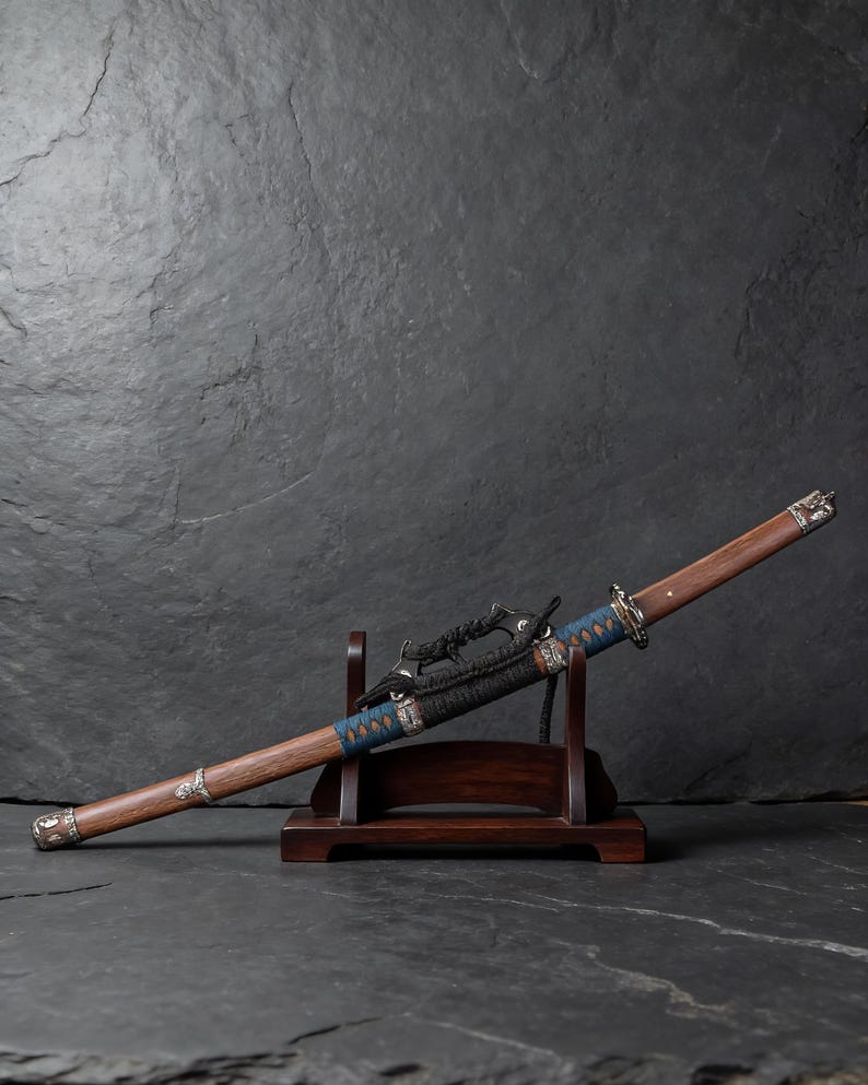 May include: A Japanese katana sword with a wooden handle and scabbard, displayed on a wooden stand. The blade is partially visible, with a blue and black wrapped hilt. Silver-coloured metal accents adorn the handle and scabbard.