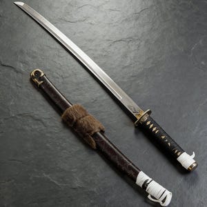 Puede incluir: Una espada katana japonesa con una hoja plateada y un mango negro, junto a su vaina marrón. La vaina tiene una envoltura de piel y detalles de cordón blanco. El mango de la espada está envuelto en un cordón negro con detalles en forma de diamante.