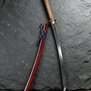 Puede incluir: Una katana japonesa con una hoja de acero pulido y un mango envuelto en marrón y blanco. La vaina es de color rojo oscuro con un cordón azul. La espada se exhibe sobre un fondo de piedra gris oscuro.