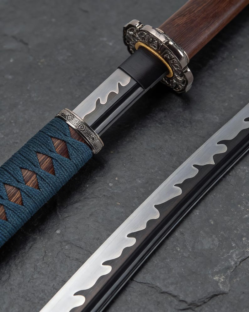 May include: Close-up of a Japanese katana sword. The blade has a wavy pattern, and the handle is wrapped in blue cord. The guard and pommel are ornate metal. The wooden handle is a dark brown colour.