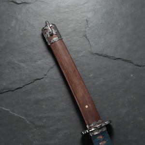 May include: A Japanese katana with a wooden handle and a silver-coloured metal guard. The handle is wrapped in blue fabric. The scabbard is a dark brown colour. The top of the scabbard has a silver-coloured metal cap with a dragon design.
