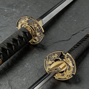 Pode incluir: Imagem detalhada de uma katana japonesa. O cabo preto apresenta um design entrançado, e a guarda dourada mostra motivos de dragões. A lâmina prateada e afiada reflete a luz.
