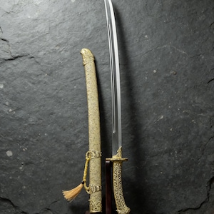May include: A decorative sword with a polished blade and ornate gold-colored handle and scabbard. The scabbard is tan with gold detailing and a tassel. The sword is displayed on a dark wooden stand.