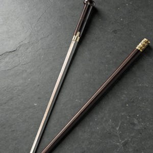 May include: A dark brown walking stick with a hidden blade. The handle and blade sheath are dark brown with gold-colored accents. The blade is silver and partially exposed. The walking stick is lying on a dark gray surface.
