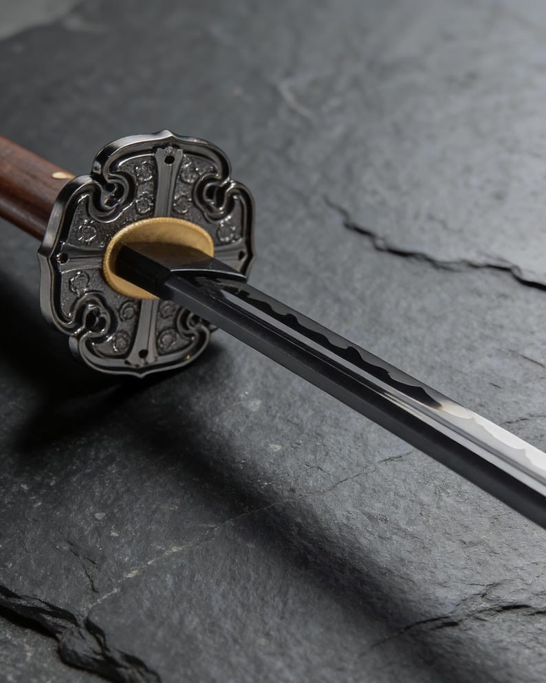 May include: Close-up of a katana sword. The sword features a dark blade, a decorative silver guard, and a wooden handle. The guard has an ornate design with a gold accent. The sword rests on a dark, textured surface.