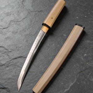 May include: A Japanese tanto knife with a light wood handle and sheath. The silver blade has a decorative pattern. The knife is partially out of its sheath, resting on a dark grey surface.