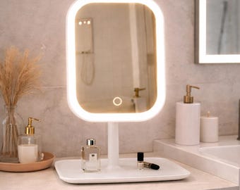 May include: A rectangular vanity mirror with a white frame and integrated LED lighting, featuring a touch-sensitive power button on the glass. The mirror is mounted on a central stand attached to a flat, rectangular white base tray.