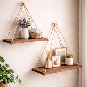 May include: Two rustic, hanging wooden shelves with natural rope supports. The shelves are stained a dark brown and hold decorative items like a vase of lavender, a woven basket with rolled towels, framed pictures, and a candle in a glass jar. The shelves are mounted on a light-colored wall.