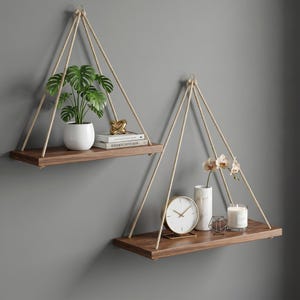 May include: Two hanging wooden shelves with rope supports. One shelf holds a potted plant, books, and a gold sculpture. The other shelf displays a clock, vase with flowers, candle, and geometric decor. The shelves are against a gray wall.