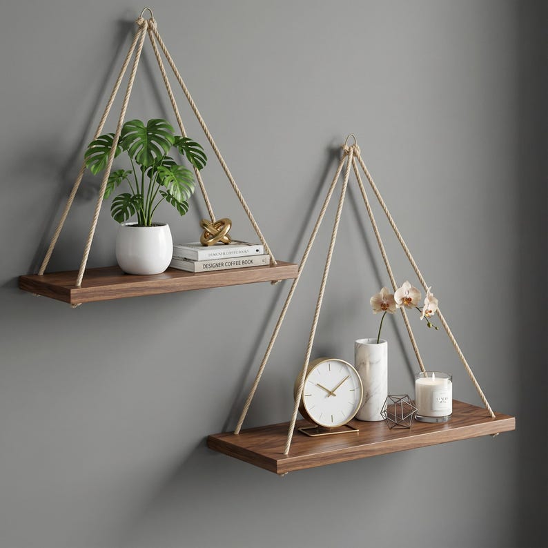 May include: Two hanging wooden shelves with rope supports. One shelf holds a potted plant, books, and a gold sculpture. The other shelf displays a clock, vase with flowers, candle, and geometric decor. The shelves are against a gray wall.