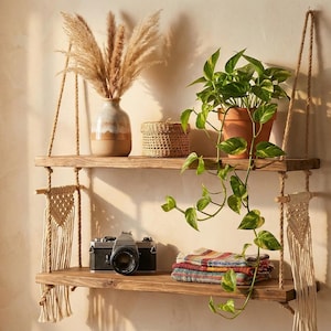 May include: A two-tiered wooden hanging shelf with macrame accents, holding a vase of dried pampas grass, a small woven basket, and a potted plant. A vintage camera and folded textiles are on the lower shelf.