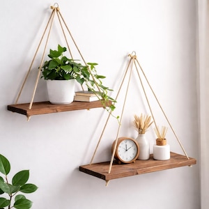 May include: Two hanging wooden shelves with plants and decorative items. The shelves are suspended by rope and attached to the wall. One shelf holds a potted plant and books, while the other has a clock, vase, and reed diffuser.