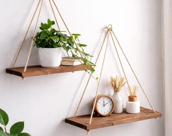 May include: Two hanging wooden shelves with plants and decorative items. The shelves are suspended by rope and attached to the wall. One shelf holds a potted plant and books, while the other has a clock, vase, and reed diffuser.