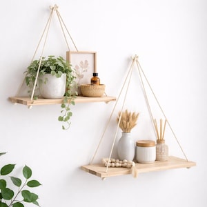 May include: Two wooden hanging shelves with natural rope supports. One shelf holds a potted plant, a framed picture, and a small bowl. The other shelf displays a vase, a jar, and decorative items. The shelves are mounted on a white wall.