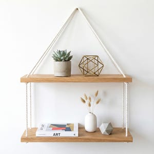 May include: A two-tiered wooden hanging shelf with white rope supports. The top shelf holds a potted succulent and a geometric gold-colored object. The bottom shelf displays a book, a white vase with dried flowers, and a geometric stone object.