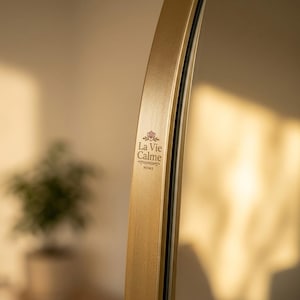 May include: A curved, gold-toned metal mirror frame featuring a smooth, brushed finish. The text La Vie Calme HOME is printed in black on the side of the frame, accompanied by a small floral icon above the brand name.