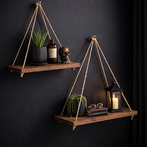 May include: Two hanging wooden shelves with rope supports against a dark wall. Each shelf holds decorative items: a potted plant, a skull, a bottle, a lantern with a lit candle, books, and eyeglasses. The shelves are made of dark wood.