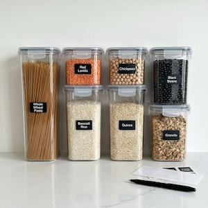 Può includere: Contenitori trasparenti in plastica per la conservazione degli alimenti riempiti con vari prodotti secchi, tra cui pasta, lenticchie, riso e granola. Ogni contenitore ha un'etichetta nera con il nome del cibo. I contenitori sono disposti ordinatamente su un piano di lavoro bianco.