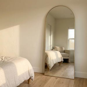 Pode incluir: Um espelho de chão alto com um topo em arco arredondado e uma moldura metálica fina. O espelho está posicionado contra uma parede lisa e reflete o interior de um quarto, incluindo uma cama com roupa de cama clara e uma cadeira perto de uma janela.