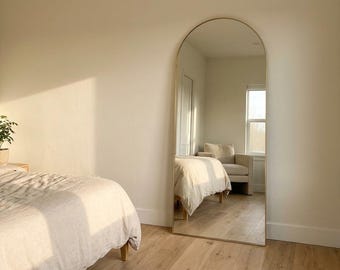May include: A tall, floor-standing mirror with a rounded arch top and a thin metallic frame. The mirror is positioned vertically against a wall, reflecting the interior of a room including a bed, a chair, and a window with natural light.