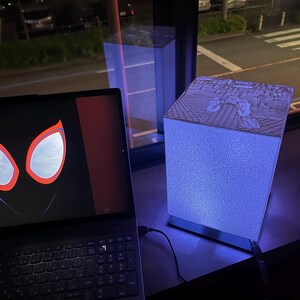 May include: A laptop displaying a graphic of red and white spider-like eyes on a black background next to a square, illuminated lamp. The lamp features a textured, light-blue glowing body and a top panel with a detailed black and white comic illustration.