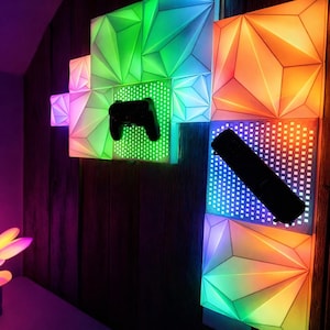 Op de afbeelding: Geometrische wandpanelen met LED-verlichting in verschillende kleuren, waaronder groen, oranje en blauw. Een zwarte gamecontroller en een zwarte afstandsbediening zijn op de panelen gemonteerd. Een decoratieve lamp met een plantachtig ontwerp is ook zichtbaar.