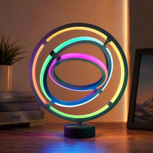 Op de afbeelding: Een cirkelvormig neonlichtarmatuur met drie in elkaar grijpende ringen die oplichten in levendige tinten geel, groen, blauw en roze. De structuur is gemonteerd op een stevige zwarte cilindrische voet, wat zorgt voor een multidimensionale lichtweergave.