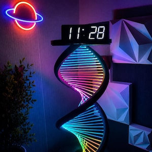 Puede incluir: Una lámpara con estructura de doble hélice negra que presenta una vibrante pantalla de luz LED multicolor que transita por un espectro de colores. Un reloj digital que muestra las 11:28 está montado sobre el marco en espiral iluminado.