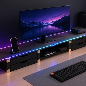 Puede incluir: Un soporte para monitor de madera oscura con cajones negros y detalles dorados, que cuenta con tiras de luz LED integradas en los bordes. El soporte sostiene un monitor de computadora ancho, un teléfono inteligente y auriculares.