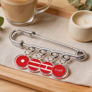 May include: A large silver safety pin with four red and white circular charms. One charm has a white dot, two have stripes, and one says "SUGA". The pin and charms are on a wooden tray with a cup of coffee and a small candle.