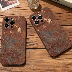 May include: Two rectangular smartphone cases featuring a textured, weathered surface with a rust-colored and dark gray patina. Each case has a cutout for a triple-lens camera system and a flash, displaying a realistic, aged metal appearance.