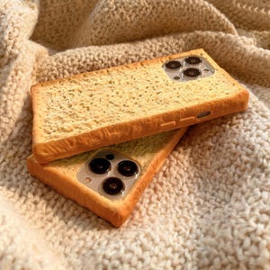 May include: Two rectangular phone cases designed to resemble slices of toasted bread. The cases feature a textured, porous surface in shades of light tan and golden brown, with cutouts for the camera lenses and side buttons on the edges.