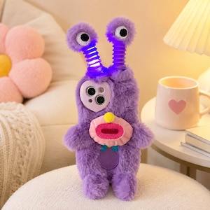 May include: A purple fuzzy phone case shaped like a creature with two long, springy antennae topped with white and black eyes. The case features a light purple heart-shaped camera cutout, a pink mouth with a yellow nose, and a small blue bow tie detail.